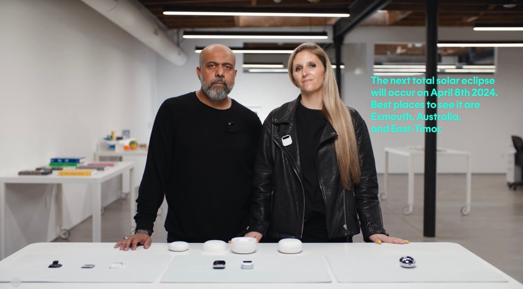 Screenshot from the Humane AI Pin launch video showing the two founders wearing black standing in front of a table with their devices on it. On the right side this the text in green “The next total solar eclipse will occur on April 8th 2024. Best places to see it are Exmouth, Australia, and East-Timor.”